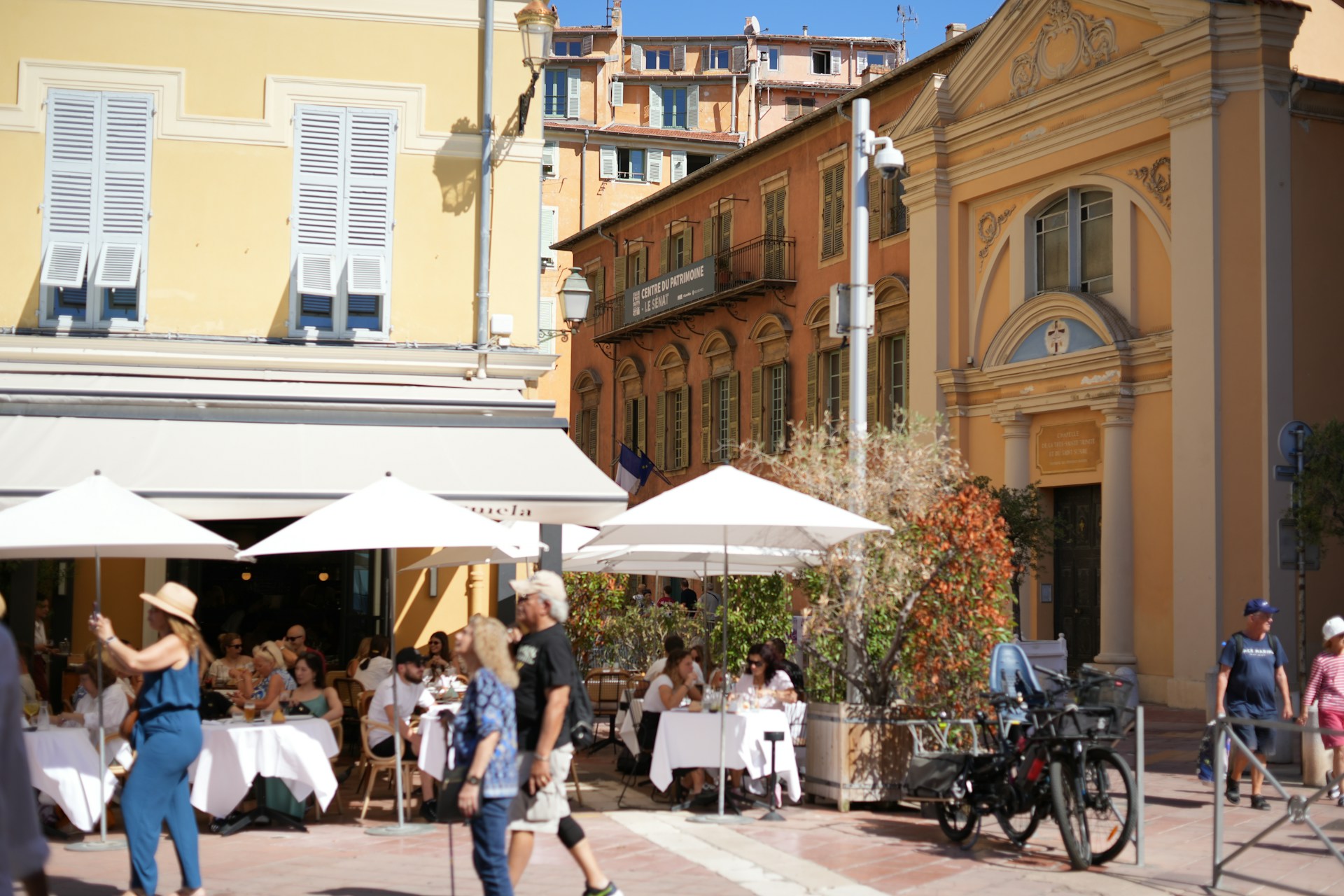 Terrasse ensoleillée sur une place à Nice, avec des façades colorées et des passants.